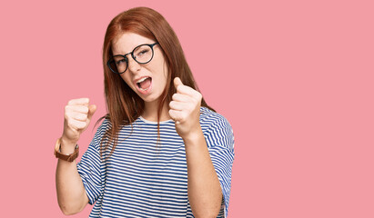 Young read head woman wearing casual clothes and glasses angry and mad raising fists frustrated and furious while shouting with anger. rage and aggressive concept.