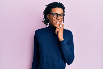 Fototapeta premium Young african american man wearing turtleneck sweater and glasses smiling looking confident at the camera with crossed arms and hand on chin. thinking positive.