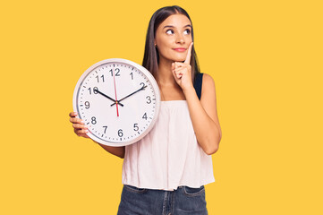 Young hispanic woman holding big clock serious face thinking about question with hand on chin, thoughtful about confusing idea