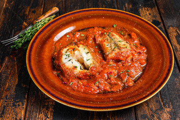 Roast hake white fish fillet with tomato sauce in a plate. Dark background. Top view