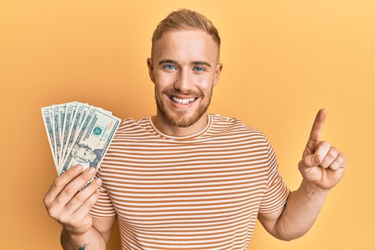 Young caucasian man holding dollars smiling with an idea or question pointing finger with happy face, number one