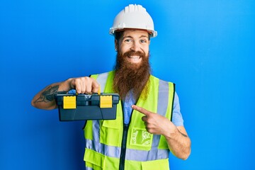 Redhead man with long beard wearing safety helmet and reflective jacket holding toolbox smiling happy pointing with hand and finger