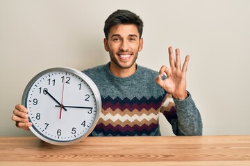 Young handsome man holding big clock doing ok sign with fingers, smiling friendly gesturing...