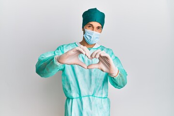 Young handsome man wearing surgeon uniform and medical mask smiling in love doing heart symbol shape with hands. romantic concept.