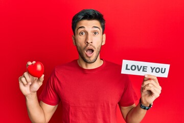 Young handsome man holding red heart and love you message afraid and shocked with surprise and amazed expression, fear and excited face.