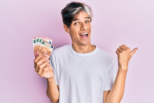 Young hispanic man holding bunch of 10 euro banknotes pointing thumb up to the side smiling happy with open mouth