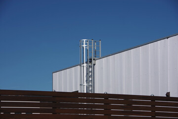 View of a segment of an industrial warehouse building under a bright blue sky with corrugated metal siding and a stainless steel access ladder to the roof and a modern wooden fence in front