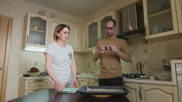 Male chef man and his wife takes photo of gingerbread cookies on table at kitchen