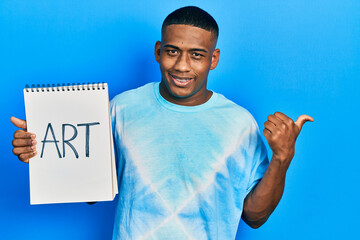 Young black man holding art notebook pointing thumb up to the side smiling happy with open mouth