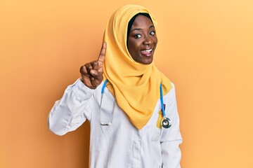 Beautiful african young woman wearing doctor uniform and hijab pointing finger up with successful idea. exited and happy. number one.