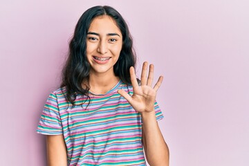 Hispanic teenager girl with dental braces wearing casual clothes showing and pointing up with fingers number five while smiling confident and happy.