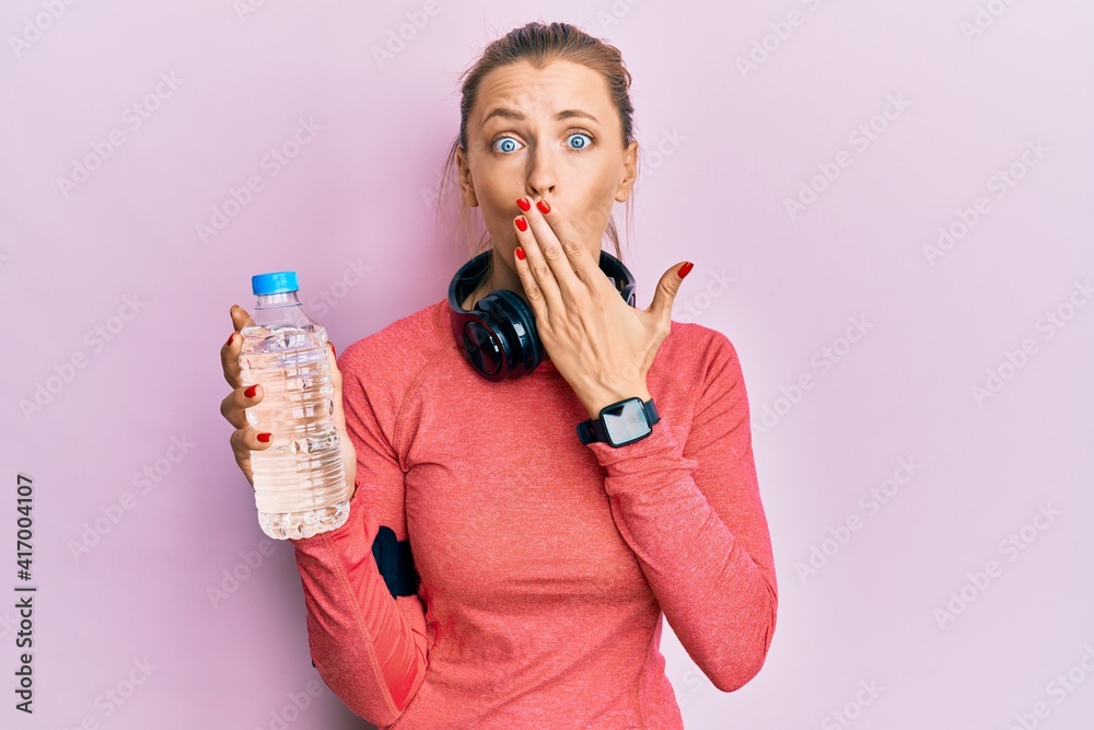 Poster beautiful caucasian sports woman wearing sportswear drinking bottle of water covering mouth with han