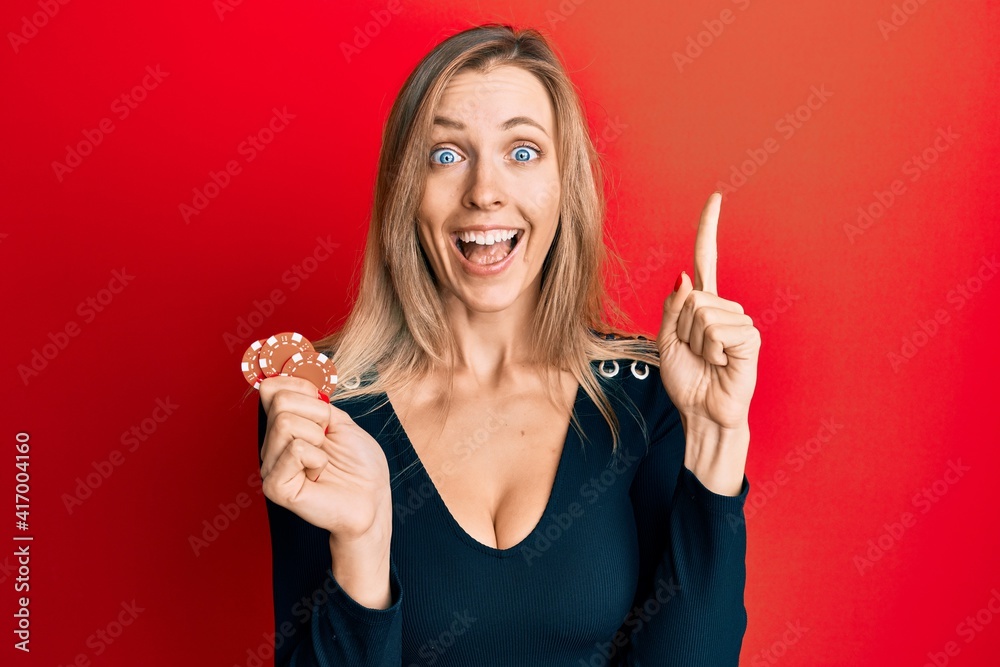 Poster Beautiful caucasian woman playing poker holding casino chips smiling with an idea or question pointing finger with happy face, number one