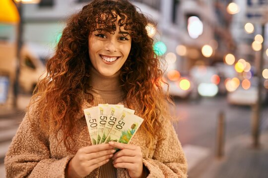 Young hispanic woman smiling happy holding israel shekels banknotes at the city