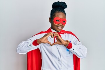 Young african american woman wearing doctor uniform and super hero costume smiling in love doing heart symbol shape with hands. romantic concept.