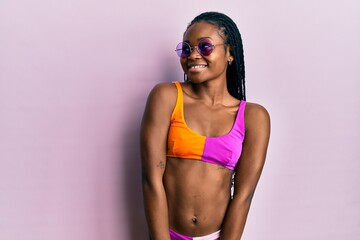 Naklejka premium Young african american woman wearing bikini and sunglasses smiling looking to the side and staring away thinking.