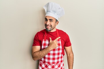 Young hispanic man wearing baker uniform cheerful with a smile of face pointing with hand and finger up to the side with happy and natural expression on face