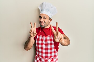 Young hispanic man wearing baker uniform smiling looking to the camera showing fingers doing victory sign. number two.