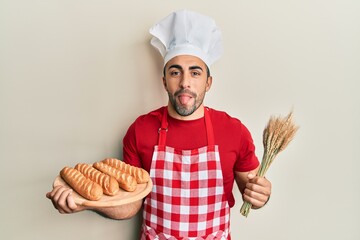 Young hispanic man wearing baker uniform holding homemade bread and spike wheat sticking tongue out happy with funny expression.