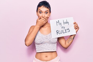 Young woman holding my body my rules banner covering mouth with hand, shocked and afraid for...
