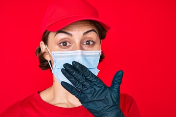 Young hispanic girl wearing delivery uniform and covid-19 safety mask covering mouth with hand, shocked and afraid for mistake. surprised expression