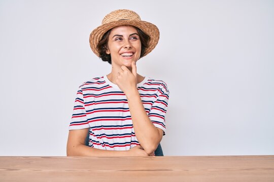Young hispanic woman wearing casual clothes and hat sitting on the table with hand on chin thinking about question, pensive expression. smiling with thoughtful face. doubt concept.