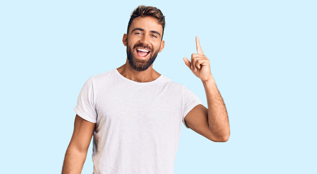 Young hispanic man wearing casual white tshirt pointing finger up with successful idea. exited and happy. number one.