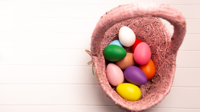 Flat lay set of decorated hand painted colorful eggs in wooden wicker rounded basket on white desk table background. Happy Easter concept composition. Image with copy free space for text message
