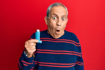 Senior hispanic man holding medical asthma inhaler scared and amazed with open mouth for surprise, disbelief face - obrazy, fototapety, plakaty