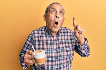Handsome senior man with grey hair drinking a cup coffee amazed and surprised looking up and pointing with fingers and raised arms.