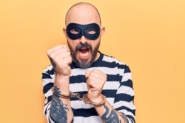 Young handsome man wearing burglar mask and handcuffs annoyed and frustrated shouting with anger, yelling crazy with anger and hand raised