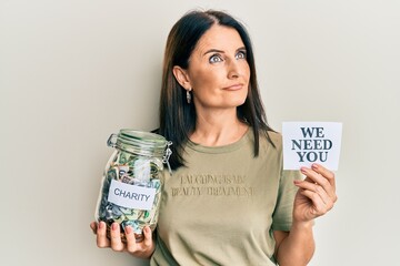 Middle age brunette woman holding charity jar with money and we need you paper smiling looking to the side and staring away thinking.