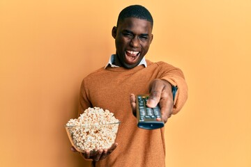 Young african american man holding television remote control eating popcorn smiling and laughing...