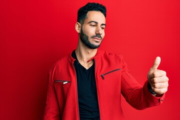 Young hispanic man wearing red leather jacket looking proud, smiling doing thumbs up gesture to the side