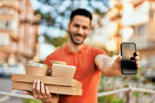 Young hispanic man showing smartphone holding take away food at the city.