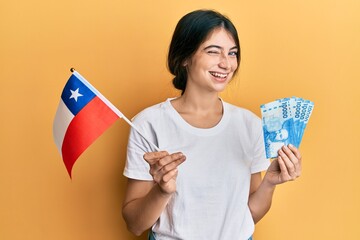 Young caucasian woman holding chile flag and chilean pesos banknotes winking looking at the camera...