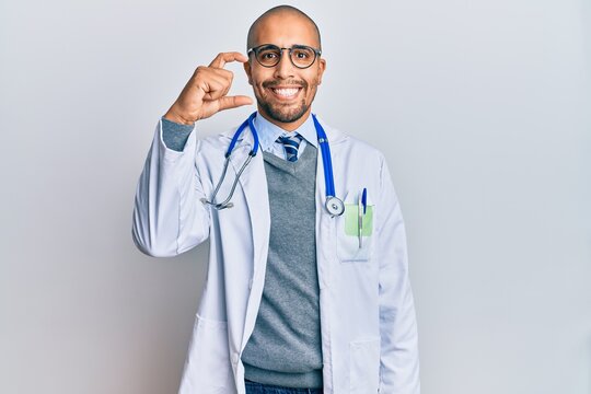 Hispanic adult man wearing doctor uniform and stethoscope smiling and confident gesturing with hand doing small size sign with fingers looking and the camera. measure concept.