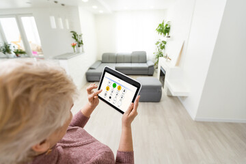 Senior woman at home standing at kitchen holding digital tablet controlling smart home system back...