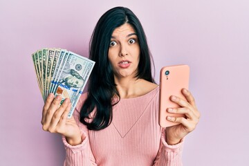 Young hispanic woman using smartphone holding united states dollar banknotes in shock face, looking skeptical and sarcastic, surprised with open mouth