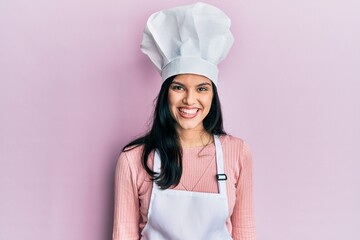 Young hispanic woman wearing baker uniform and cook hat looking positive and happy standing and smiling with a confident smile showing teeth