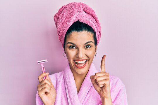 Young hispanic woman wearing shower bathrobe holding razor smiling with an idea or question pointing finger with happy face, number one
