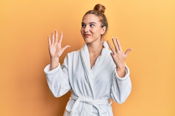 Young blonde woman wearing bathrobe showing and pointing up with fingers number nine while smiling...