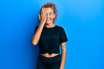 Beautiful caucasian teenager girl wearing black sportswear covering one eye with hand, confident smile on face and surprise emotion.