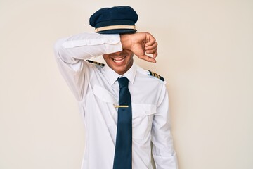 Young hispanic man wearing airplane pilot uniform covering eyes with arm smiling cheerful and funny. blind concept.