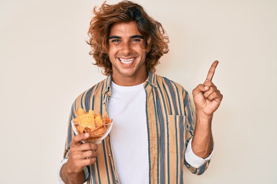 Young hispanic man holding nachos potato chips smiling happy pointing with hand and finger to the side