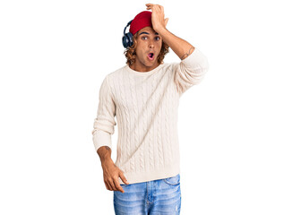 Young hispanic man listening to music using headphones surprised with hand on head for mistake, remember error. forgot, bad memory concept.