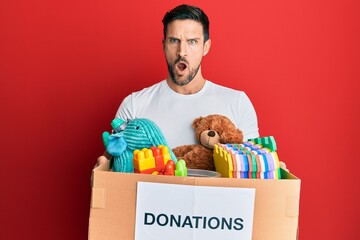Young handsome man holding donation box with toys in shock face, looking skeptical and sarcastic,...