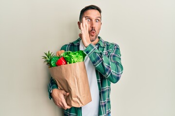 Handsome man with beard holding paper bag with groceries hand on mouth telling secret rumor, whispering malicious talk conversation