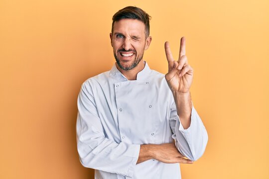 Handsome man with beard wearing professional cook uniform smiling with happy face winking at the camera doing victory sign. number two.