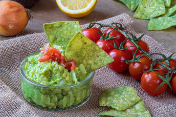 Fresh homemade guacamole dip with green, avocado flavored, tortilla chips. Gluten free traditional Mexican cuisine or dish. Cherry tomatoes, onion and lemon in background on brown rustic cloth.